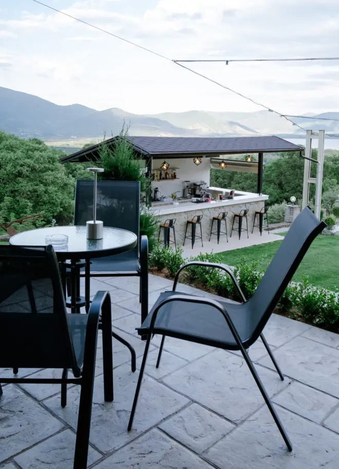 realistic photo of an outdoor setting featuring a small glass round table with chairs and a bar in the background around a lush greenery and the lake