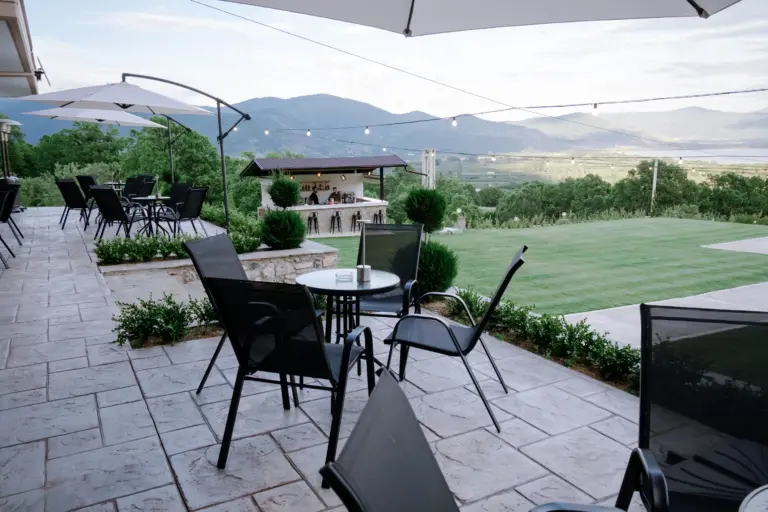 realistic photo of the hotel main balcony area with a lot of coffee tables with chairs and large umbrellas over them and a view of the outdoor bar in the background